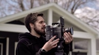 A person is outdoors holding a professional RED camera, appearing focused while adjusting the equipment. The background shows parts of a house and blurred trees, suggesting a calm and natural setting.