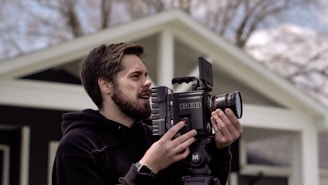 A person is outdoors holding a professional RED camera, appearing focused while adjusting the equipment. The background shows parts of a house and blurred trees, suggesting a calm and natural setting.