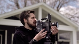 A person is outdoors holding a professional RED camera, appearing focused while adjusting the equipment. The background shows parts of a house and blurred trees, suggesting a calm and natural setting.