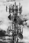 A large telecommunications tower is equipped with numerous antennas and satellite dishes. The structure is complex, with cables and metal components extending outward, set against a background of partly cloudy skies.