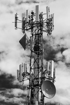 A large telecommunications tower is equipped with numerous antennas and satellite dishes. The structure is complex, with cables and metal components extending outward, set against a background of partly cloudy skies.