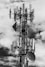 A large telecommunications tower is equipped with numerous antennas and satellite dishes. The structure is complex, with cables and metal components extending outward, set against a background of partly cloudy skies.