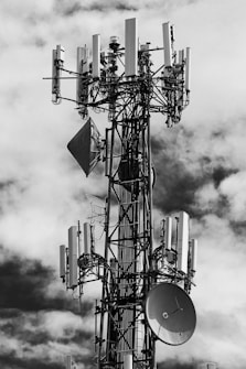 A large telecommunications tower is equipped with numerous antennas and satellite dishes. The structure is complex, with cables and metal components extending outward, set against a background of partly cloudy skies.