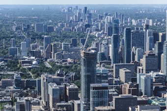 The image captures a panoramic view of a modern cityscape with numerous high-rise buildings and skyscrapers. The foreground features a dense cluster of buildings, with a mix of glass and concrete structures, while the background extends into a lush, green area, suggesting urban planning that incorporates nature.