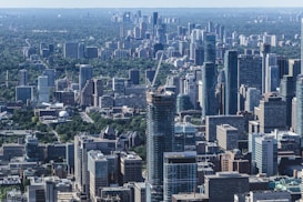 The image captures a panoramic view of a modern cityscape with numerous high-rise buildings and skyscrapers. The foreground features a dense cluster of buildings, with a mix of glass and concrete structures, while the background extends into a lush, green area, suggesting urban planning that incorporates nature.