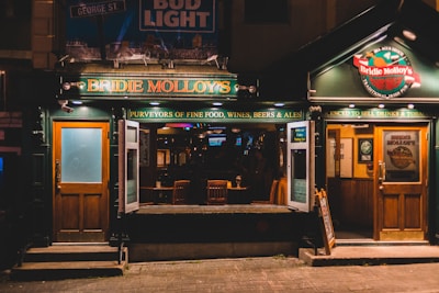 A cozy Irish pub with wooden doors and the inviting glow of warm lighting. The establishment's name, Bridie Molloy's, is prominently displayed, suggesting a traditional and welcoming ambiance. Inside, the dimly lit interior with wooden furniture and a glass of beer on a table can be seen, offering a classic pub experience.