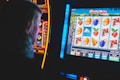 A person is closely observing a digital slot machine with colorful fruit symbols, bars, and a bell. The slot machine screen displays symbols like oranges, plums, cherries, and clouds. The background is dimly lit, creating a focused and somewhat intense atmosphere.