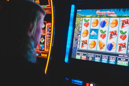 A person is closely observing a digital slot machine with colorful fruit symbols, bars, and a bell. The slot machine screen displays symbols like oranges, plums, cherries, and clouds. The background is dimly lit, creating a focused and somewhat intense atmosphere.