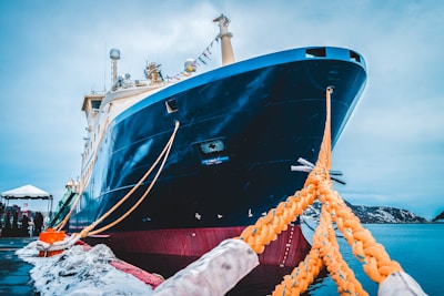 A large ship is docked at a port, secured with thick yellow ropes. The ship's hull is predominantly dark blue with a white upper structure. Snow can be seen on the ground at the dock, indicating a cold environment. In the background, there are mountains partially covered in snow, and the sky is overcast. The ship appears to be equipped with various communication antennas and a large forward deck.