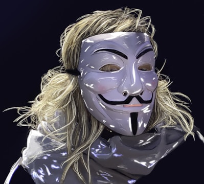 A person wears a reflective, stylized mask resembling the iconic Guy Fawkes design. Long, light-colored hair flows around the mask, and the background is a dark, solid color. The mask features distinct black markings with a smooth surface.