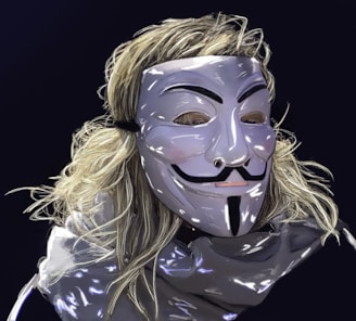 A person wears a reflective, stylized mask resembling the iconic Guy Fawkes design. Long, light-colored hair flows around the mask, and the background is a dark, solid color. The mask features distinct black markings with a smooth surface.