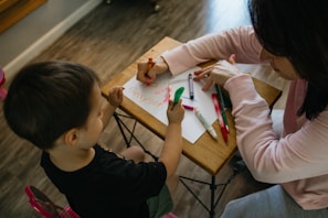 A young child and an adult are sitting at a small table engaged in a drawing activity. Various colored crayons and markers are scattered on the table surface, and the adult is assisting the child in writing or drawing. The setting appears to be indoors with wooden flooring.