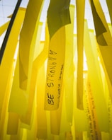 Yellow ribbons with motivational messages hang vertically. One prominent ribbon displays the words 'Be Strong' along with hearts. The background consists of similar yellow ribbons, creating a layered effect.