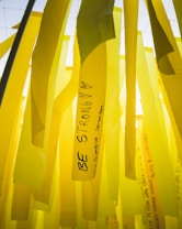 Yellow ribbons with motivational messages hang vertically. One prominent ribbon displays the words 'Be Strong' along with hearts. The background consists of similar yellow ribbons, creating a layered effect.