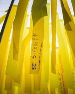 Yellow ribbons with motivational messages hang vertically. One prominent ribbon displays the words 'Be Strong' along with hearts. The background consists of similar yellow ribbons, creating a layered effect.