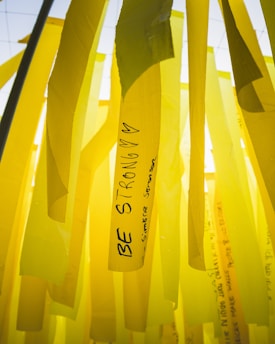 Yellow ribbons with motivational messages hang vertically. One prominent ribbon displays the words 'Be Strong' along with hearts. The background consists of similar yellow ribbons, creating a layered effect.