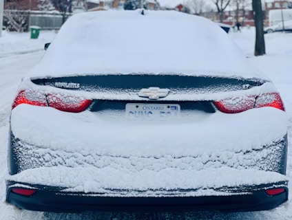 A snow-covered car parked on a residential street, with snow blanketing the rear windshield and trunk, partially obscuring the car's logo and license plate.