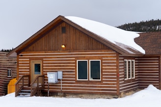 A wooden cabin is set in a snowy landscape, featuring a sloped roof partially covered with snow. The cabin has a small porch with steps leading to the entrance and a row of windows along the side. The siding consists of horizontal wooden planks with white accents between them.