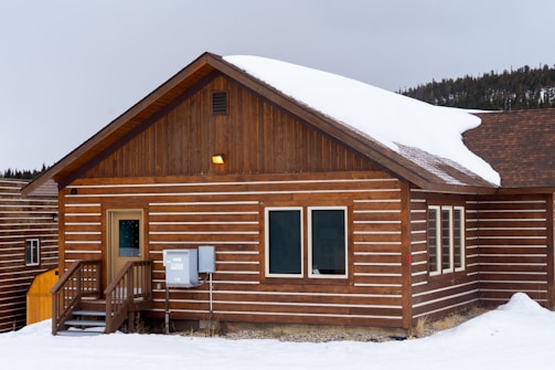 A wooden cabin is set in a snowy landscape, featuring a sloped roof partially covered with snow. The cabin has a small porch with steps leading to the entrance and a row of windows along the side. The siding consists of horizontal wooden planks with white accents between them.