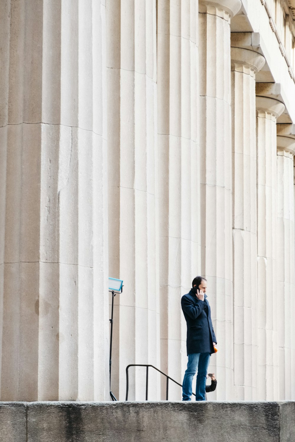 mulher na jaqueta preta e jeans azuis em pé ao lado da parede de concreto branca durante o dia