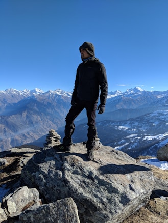 A person dressed in dark outdoor clothing stands confidently on a rocky mountain summit. The background features a breathtaking view of snow-capped mountains under a clear blue sky.