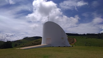 A modern white chapel with a minimalist design is situated in a grassy landscape. The structure features a large cross on top and a winding external staircase. The sky above is partly cloudy with patches of blue, and rolling green hills are visible in the background.