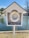 A white lifebuoy is housed in a small wooden structure resembling a miniature open-fronted house with a gable roof. It is positioned by the edge of a serene body of water, with trees visible in the background. The lifebuoy is accompanied by a neatly coiled rope placed at the bottom of the structure.