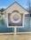 A white lifebuoy is housed in a small wooden structure resembling a miniature open-fronted house with a gable roof. It is positioned by the edge of a serene body of water, with trees visible in the background. The lifebuoy is accompanied by a neatly coiled rope placed at the bottom of the structure.