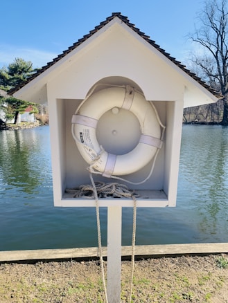 A white lifebuoy is housed in a small wooden structure resembling a miniature open-fronted house with a gable roof. It is positioned by the edge of a serene body of water, with trees visible in the background. The lifebuoy is accompanied by a neatly coiled rope placed at the bottom of the structure.