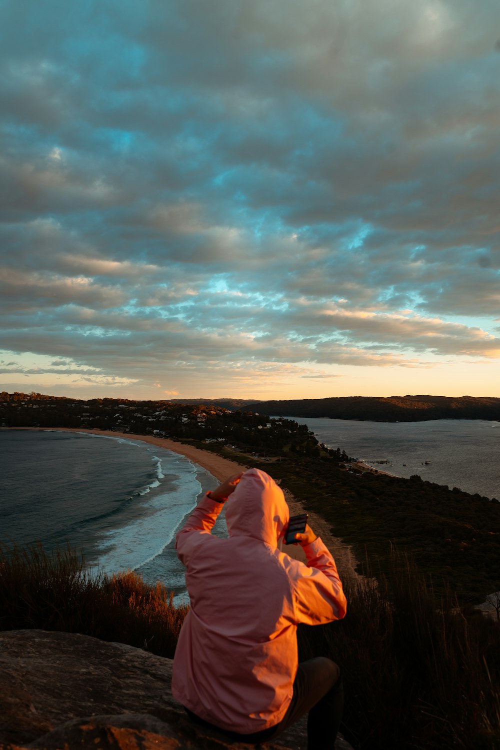 woman in pink hoodie sitting on brown rock formation near body of water during daytime