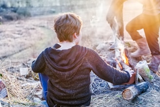 A person sitting by a campfire in an outdoor setting with dry grass and logs scattered around. The person is wearing a dark hoodie and blue pants, focusing on maintaining the fire, while another person, wearing boots, stands nearby.