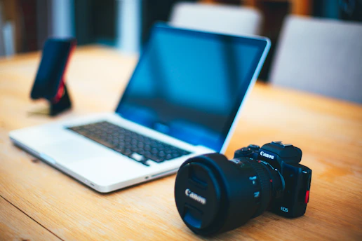 A laptop with a black keyboard sits on a wooden table alongside a digital camera with Canon branding. In the background, a smartphone is positioned upright in a stand. The setting suggests a workspace or a photographer's setup.