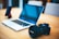 A laptop with a black keyboard sits on a wooden table alongside a digital camera with Canon branding. In the background, a smartphone is positioned upright in a stand. The setting suggests a workspace or a photographer's setup.