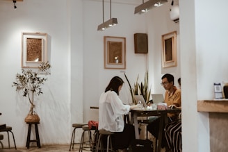 A cozy cafe interior with two people working on laptops at a shared table. The setting features minimalist decor with white walls, framed artwork, and a small indoor plant. There are several stools around the table, and the lighting is warm and inviting.