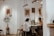 A cozy cafe interior with two people working on laptops at a shared table. The setting features minimalist decor with white walls, framed artwork, and a small indoor plant. There are several stools around the table, and the lighting is warm and inviting.