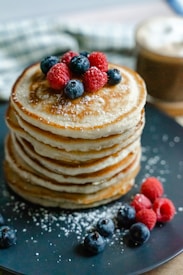 A stack of fluffy golden-brown pancakes topped with fresh raspberries and blueberries is placed on a dark plate. Light dusting of powdered sugar surrounds the plate, adding a touch of sweetness. In the background, there is a blurry hint of a checkered cloth and a cup of coffee.