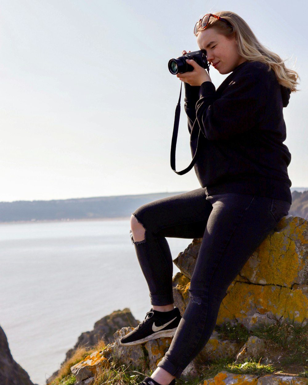 woman in black jacket and black pants sitting on rock while holding camera