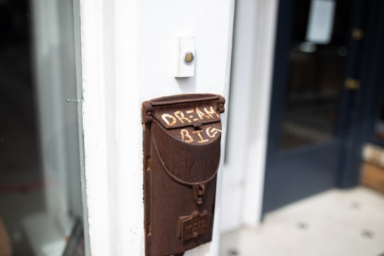 A vintage brown mailbox is attached to a white wall. The mailbox has the words 'DREAM BIG' painted on it in white letters. The setting appears to be outside a building with glass windows and a dark door visible in the background.