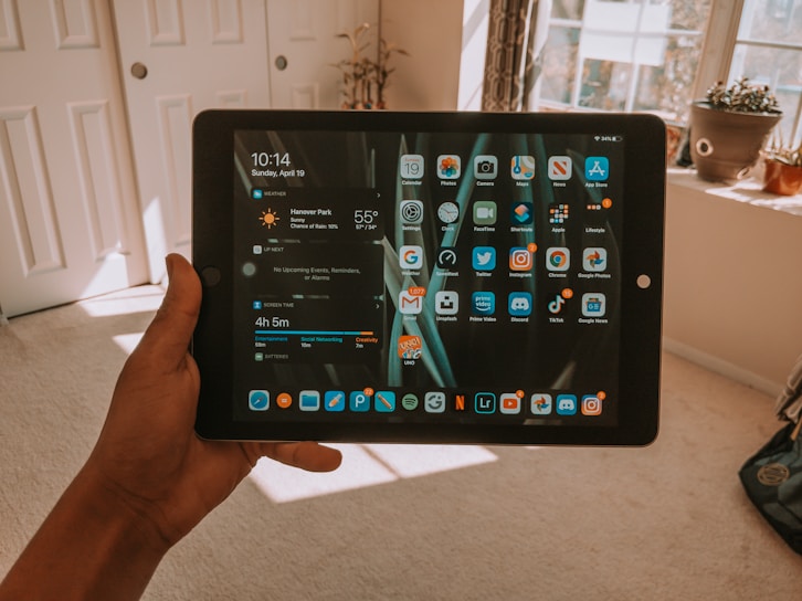 A hand holding a tablet displaying a home screen with various app icons and notifications. The screen shows details such as time, weather, and screen time. The background is a bright room with light streaming in from the window and potted plants on the windowsill.