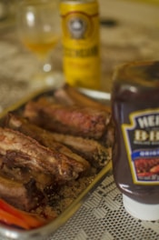 A close-up view of a table with a tray of cooked ribs, a bottle of barbecue sauce, and a can of beer. There is also a glass with an orange-colored beverage in the background. The tablecloth is white and patterned.