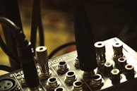 A close-up view of an audio interface or mixer with various input jacks and connected cables. The surface is metallic and displays text and symbols indicating different functionalities. The lighting is dim and the focus is on the central area of the device.