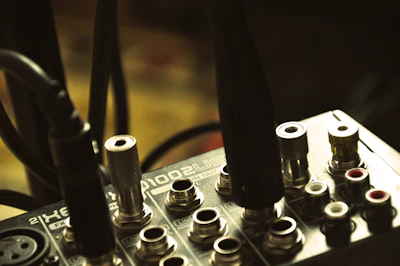 A close-up view of an audio interface or mixer with various input jacks and connected cables. The surface is metallic and displays text and symbols indicating different functionalities. The lighting is dim and the focus is on the central area of the device.