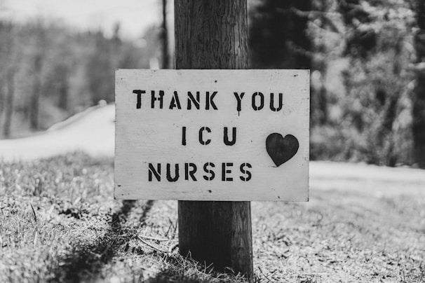 A black and white photo of a wooden sign attached to a pole with the message 'THANK YOU ICU NURSES' alongside a heart symbol. The sign is positioned outdoors with a blurred background of trees and grass.