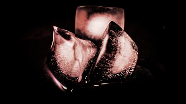 Three ice cubes, two of which are partially melted with visible cracks and bubbles, are arranged against a dark background. The lighting highlights the texture and transparency of the cubes, creating a dramatic and abstract appearance.
