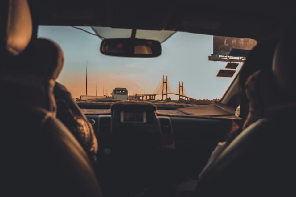 The image shows a car's interior with two people seated inside, facing each other and looking out at a bridge visible through the windshield. The car is parked on a street, and there are several buildings in the background.