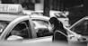 A black and white image captures a woman wearing a mask standing beside a car with a taxi sign. She carries a woven basket over her shoulder. The car window is partially open, revealing a masked driver inside. Background vehicles and cityscape elements are blurred.