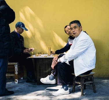 Three men are gathered around a small table playing a board game, possibly involving stacking pieces. Two men are sitting on small wooden stools, while one is standing to the left, partially out of frame. The man in the foreground wears a white jacket and sneakers, and another man beside him laughs while wearing a striped sweater. The third man, who is focused on the game, wears a blue cap and dark jacket. They are set against a yellow wall and sitting on a concrete floor, with sunlight casting shadows.