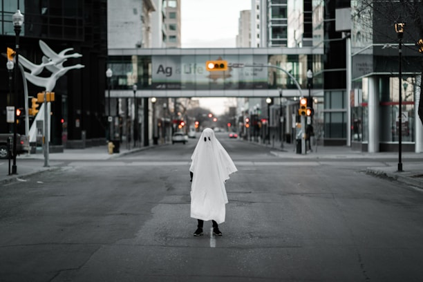 A solitary figure dressed in a simple ghost costume stands in the middle of an empty city street. The urban setting is framed by tall buildings and a pedestrian bridge in the background. The scene appears to be depicted in a cinematic style with a focus on contrast and lighting.