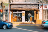 A storefront with a traditional Chinese design features a wooden facade and glass doors with decorative patterns. A blue car is parked on the street in front of the store. There is a tree with green leaves on the sidewalk and a red lantern hanging from it. The interior of the store is dimly visible, with some items on display.
