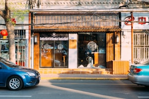 A storefront with a traditional Chinese design features a wooden facade and glass doors with decorative patterns. A blue car is parked on the street in front of the store. There is a tree with green leaves on the sidewalk and a red lantern hanging from it. The interior of the store is dimly visible, with some items on display.